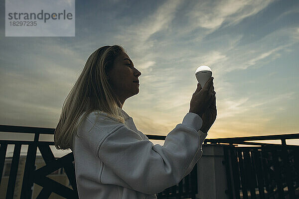 Woman holding LED bulb standing under sky at sunset