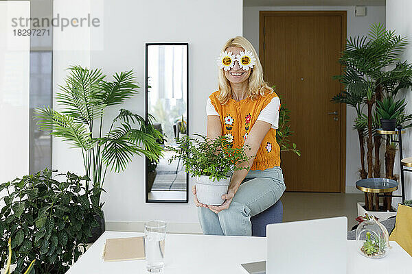 Glückliche Frau mit Blumensonnenbrille  die zu Hause auf einem Stuhl mit einer Pflanze sitzt