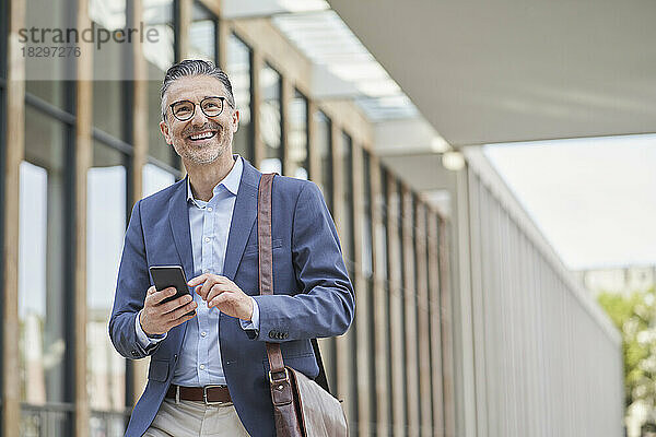 Lächelnder reifer Geschäftsmann hält Mobiltelefon vor Gebäude