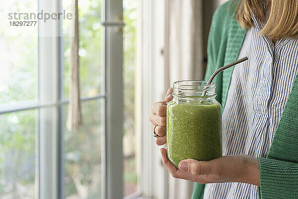 Hände einer Frau mit grünem Smoothie am Fenster