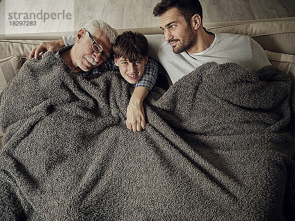 Grandfather  father and son snuggling under a blanket on couch in living room