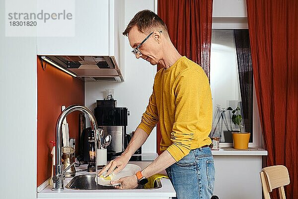 Mann mittleren Alters mit Brille beim abendlichen Geschirrspülen in der Küche