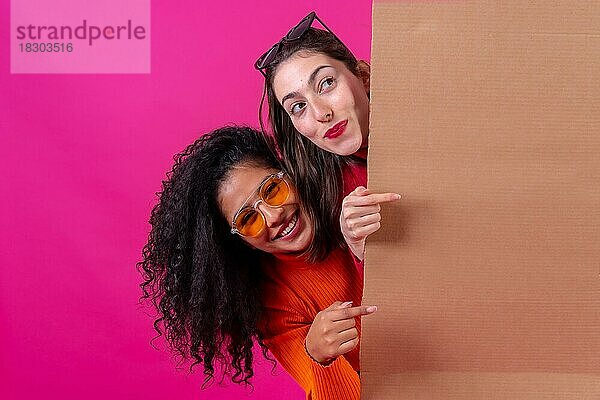 Zwei lächelnde schöne Freundinnen  die auf ein Pappschild vor einem rosa Hintergrund zeigen