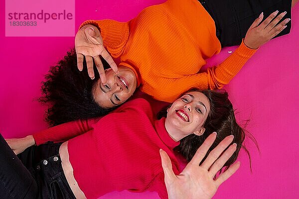 Zwei multiethnische Frauen liegen lächelnd in einer trendigen Pose vor einem rosa Hintergrund  Lifestyle