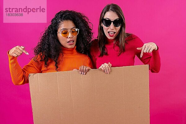 Zwei Freundinnen lächelnd und auf das Pappschild auf einem rosa Hintergrund zeigend