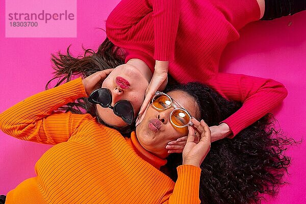 Zwei multiethnische Frauen mit Sonnenbrillen liegen lächelnd auf einem rosa Hintergrund  Lifestyle