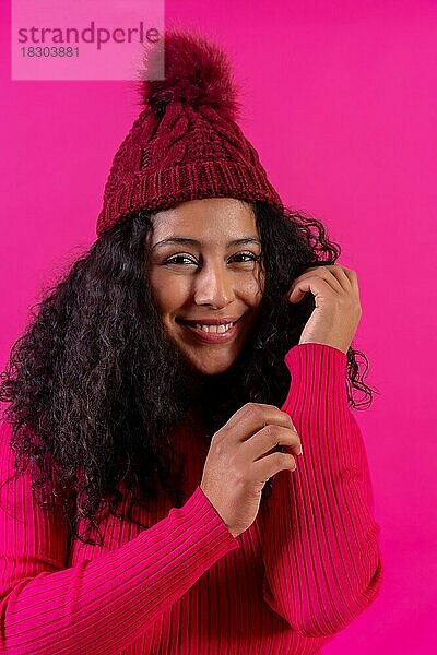 Lockenköpfige Frau mit Wollmütze auf rosa Hintergrund  lächelnd  Studioaufnahme
