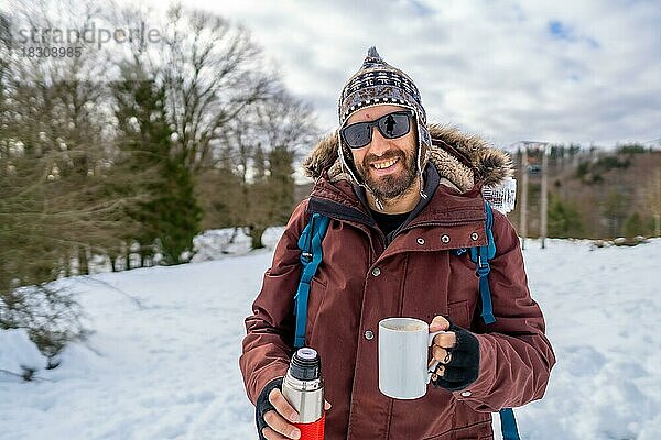 Porträt eines Mannes  der im Winter im Schnee einen heißen Kaffee aus einer Thermoskanne frühstückt
