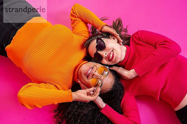 Zwei multiethnische Frauen mit Sonnenbrillen liegen lachend auf einem rosa Hintergrund  Lifestyle