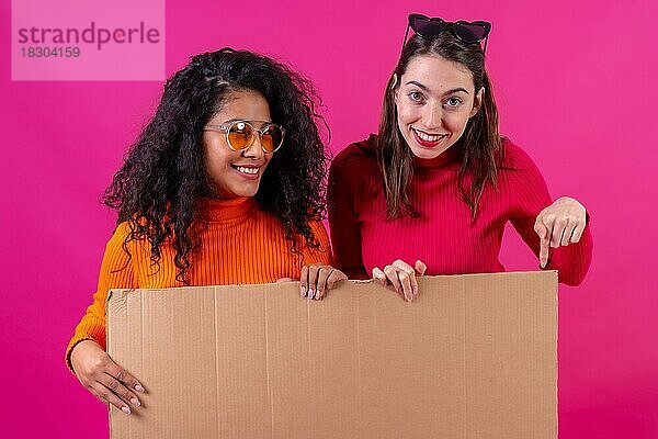 Zwei multiethnische Freundinnen  die lächelnd auf ein Pappschild vor rosa Hintergrund zeigen