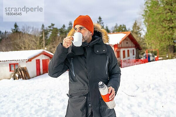 Mann trinkt Kaffee aus einer heißen Thermoskanne im Winter im Schnee neben einer Hütte