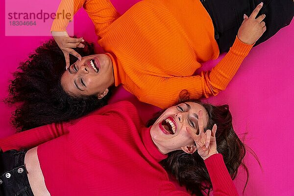 Zwei multiethnische Frauen liegen lächelnd in einer trendigen Pose vor einem rosa Hintergrund  Lifestyle