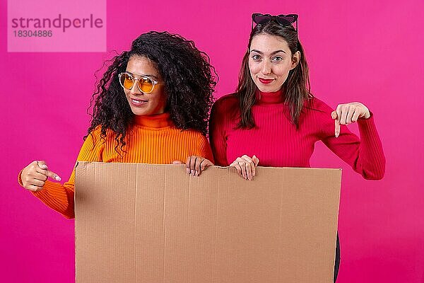 Zwei Freundinnen lächelnd und auf das Pappschild auf einem rosa Hintergrund zeigend
