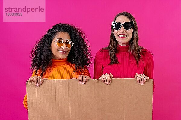 Zwei lächelnde multiethnische Freundinnen halten ein Pappschild vor einem rosa Hintergrund
