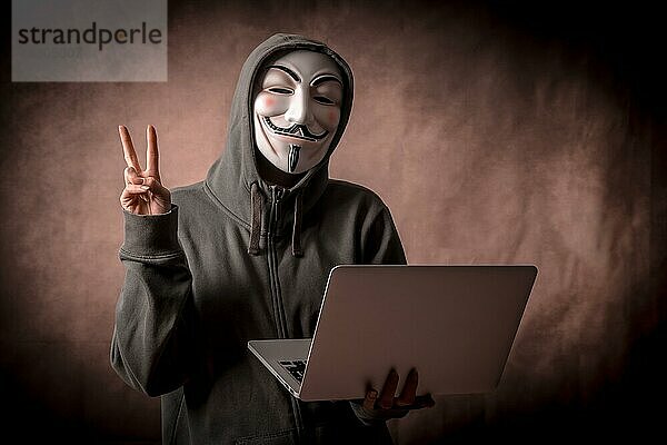 Hacker mit anonymer Maske mit einem Laptop  der das Siegeszeichen macht  Studioaufnahme
