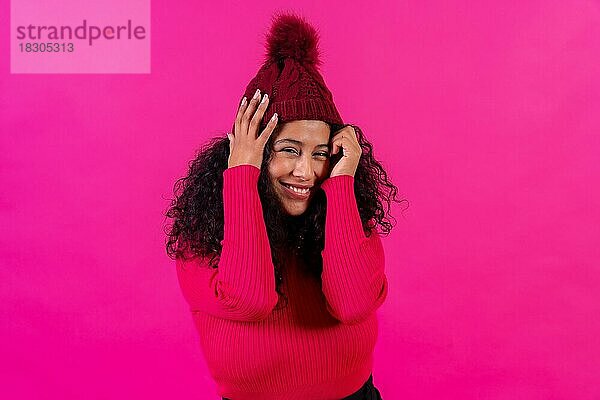 Lockenköpfige Frau mit Wollmütze vor rosa Hintergrund  schüchtern lächelnd  Studioaufnahme
