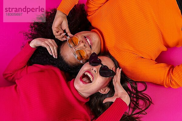 Zwei multiethnische Frauen mit Sonnenbrillen liegen lächelnd auf einem rosa Hintergrund  Lifestyle