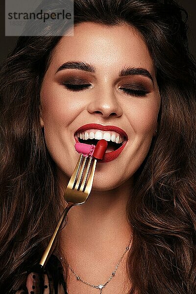 Schönes Mädchen mit roten Lippen und klassischen Make-up und Locken mit Lippenstift in der Hand. Schönes Gesicht. Foto im Studio aufgenommen