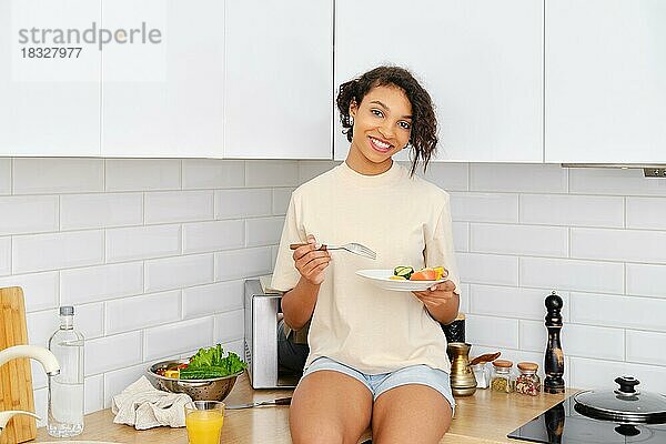 Glückliche afroamerikanische Frau sitzt in der Küche mit Teller mit Frühstück in den Händen