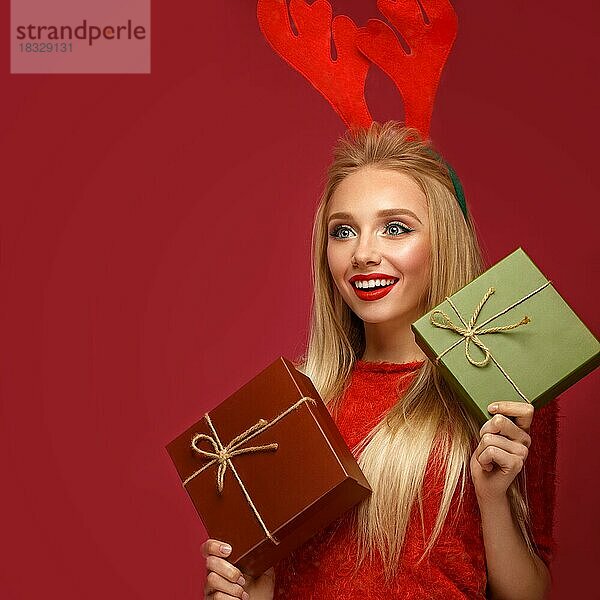 Schönes blondes Mädchen in einem Neujahrsbild mit Kisten mit Geschenken in den Händen und Hirschhörnern auf dem Kopf. Schönes Gesicht mit festlichem Make-up. Foto im Studio aufgenommen