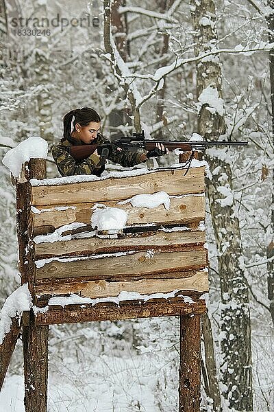 Attraktives Jägermädchen mit Gewehr auf Jagdturm im Winterwald