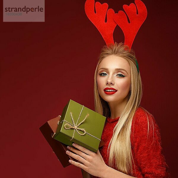 Schönes blondes Mädchen in einem Neujahrsbild mit Kisten mit Geschenken in den Händen und Hirschhörnern auf dem Kopf. Schönes Gesicht mit festlichem Make-up. Foto im Studio aufgenommen