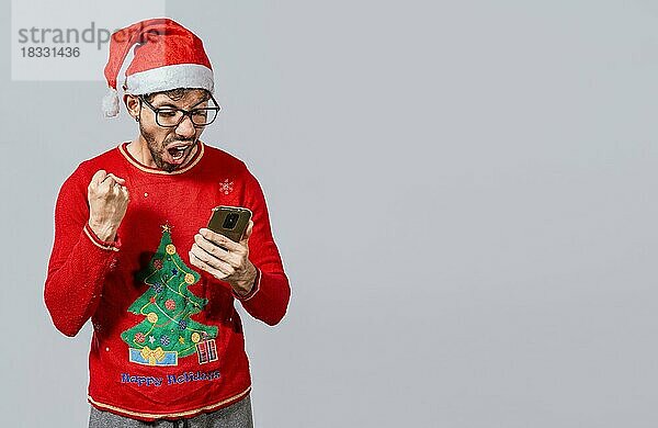 Aufgeregter Mann in Weihnachtsmütze schaut auf Handy und feiert. Menschen in santa Hut Blick auf Handy aufgeregt  aufgeregt junger Mann in Weihnachten Hut mit Handy feiern