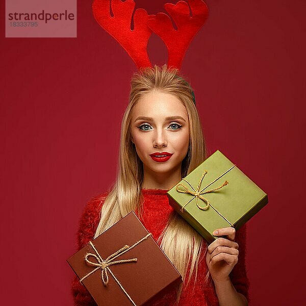 Schönes blondes Mädchen in einem Neujahrsbild mit Kisten mit Geschenken in den Händen und Hirschhörnern auf dem Kopf. Schönes Gesicht mit festlichem Make-up. Foto im Studio aufgenommen