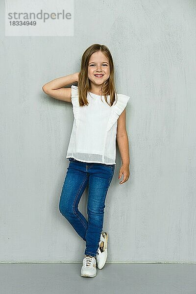 Kleines Mädchen in weißer Bluse und Jeans lehnt sich an die Wand und hält eine Hand hinter den Kopf