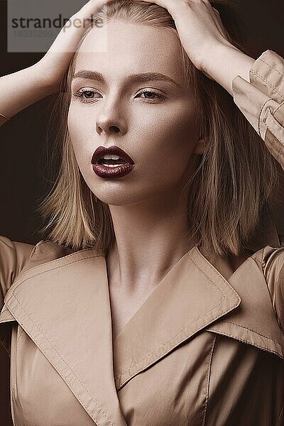 Schöne blonde Frau in einem hellen Mantel und dunklen Lippen  zeigt verschiedene Posen. Schönes Gesicht. Bild im Studio aufgenommen