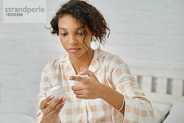Afrikanische Amerikanerin  die einen offenen Tiegel mit Feuchtigkeitscreme in der Hand hält und bereit ist  diese auf ihren Körper aufzutragen (Foto mit geringer Schärfentiefe)