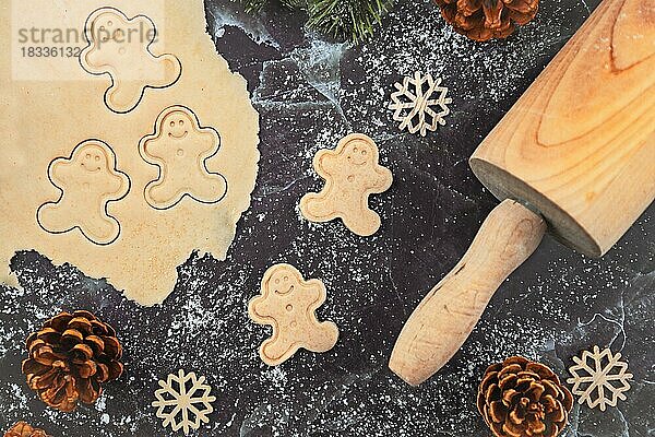 Flat Lay-Konzept zum Backen von Weihnachtsplätzchen mit ausgerolltem Plätzchenteig  Plätzchen in Form von glücklichen Lebkuchenmännern  Nudelholz und saisonalen Tannenzweigen auf dunklem Marmor-Hintergrund