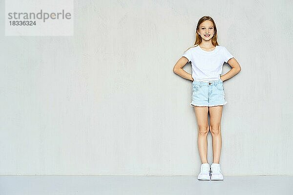 Junges sportliches Mädchen in weißem T-Shirt und Jeansshorts lehnt sich mit beiden Händen hinter dem Rücken an die Wand