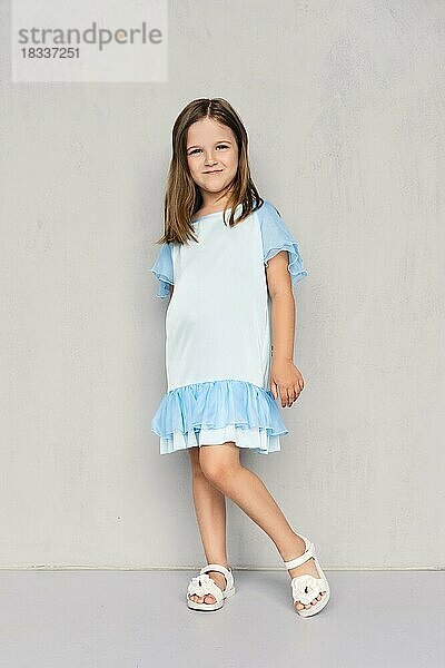 Nettes kleines Mädchen in blauem Kleid und weißen Sandalen posiert in der Nähe der grauen Wand
