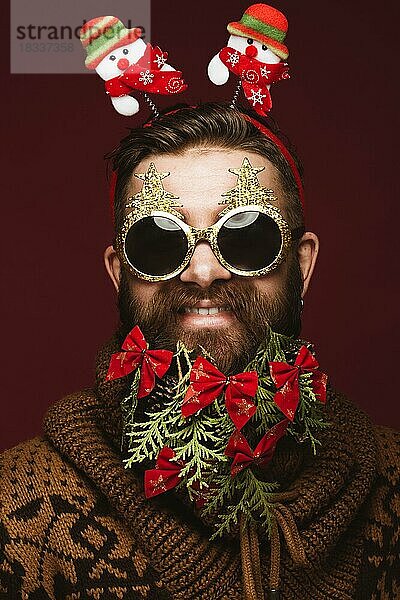 Lustiger bärtiger Mann in einem Neujahrsbild als Weihnachtsmann mit Dekorationen auf seinem Bart. Fest der Weihnacht. Fotos im Studio aufgenommen