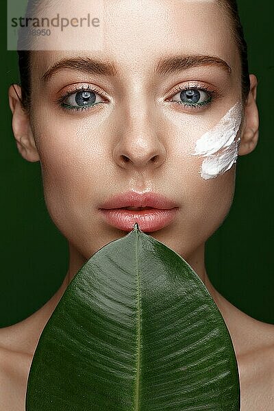 Schönes frisches Mädchen mit Kosmetikcreme auf dem Gesicht  natürliches Make-up und grüne Blätter. Schönes Gesicht. Foto im Studio aufgenommen