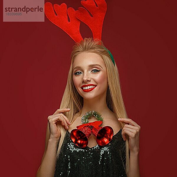 Schönes blondes Mädchen in einem Neujahrsbild mit Weihnachtsglocken um ihren Hals und Hirschhörnern auf ihrem Kopf. Schönes Gesicht mit festlichem Make-up. Foto im Studio aufgenommen