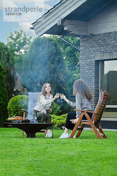 Zwei Freundinnen sitzen beim Grill im Garten und stoßen mit Sekt an