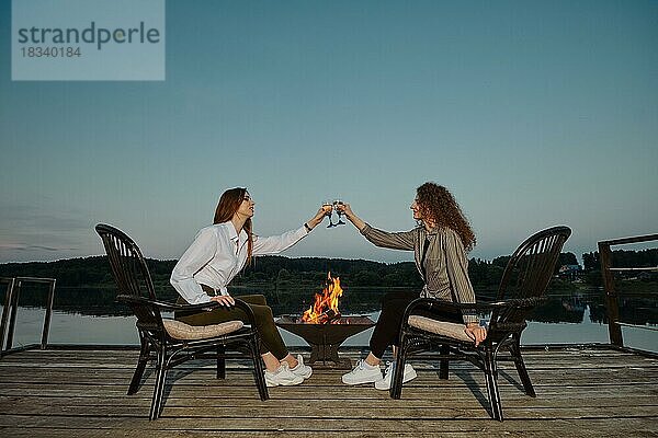 Glückliche Frauen stoßen mit Weißwein an und sitzen auf dem Pier in der Nähe des Feuers