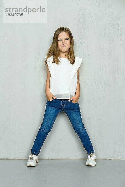 Kleines Mädchen in weißer Bluse und Jeans lehnt an der Wand  hält die Hände in den Taschen und stellt die Beine breit