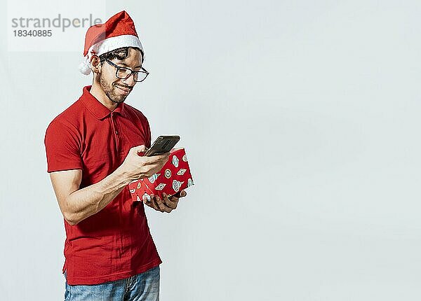 Mann mit Weihnachtsmütze hält Geschenkbox und lächelt auf Smartphone. Weihnachten Mann hält Geschenk-Box und Telefon isoliert. gutaussehender Mann mit Weihnachtsmütze  der ein Geschenk hält und auf das Telefon schaut