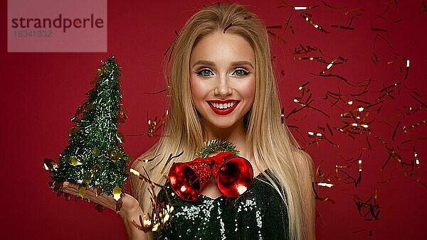 Schönes blondes Mädchen in einem Neujahrsbild mit Weihnachtsglocken um ihren Hals und grünem Baum. Schönes Gesicht mit festlichem Make-up. Foto im Studio aufgenommen