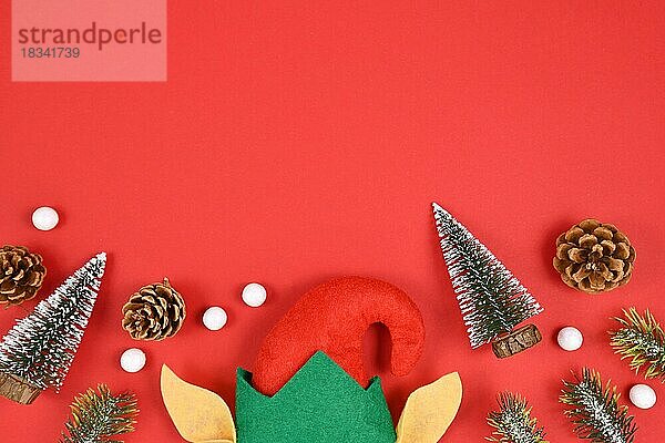 Saisonales Flatlay mit Weihnachtselfenmütze und -ohren  kleinen Tannenbäumen und -zapfen und weißen Schneebällen unten und leerem Kopierbereich auf rotem Hintergrund oben