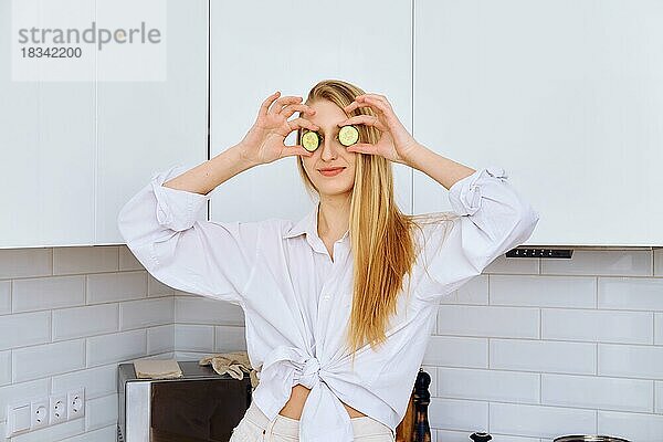 Fröhliche junge Frau hält sich beim Kochen Gurkenstücke an die Augen