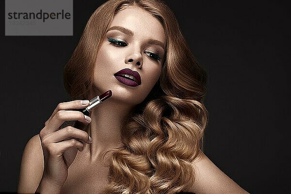 Schöne Blondine in Hollywood-Manier mit Locken  dunklen Lippen  Lippenstift in der Hand. Schönes Gesicht und Haar. Bild im Studio aufgenommen