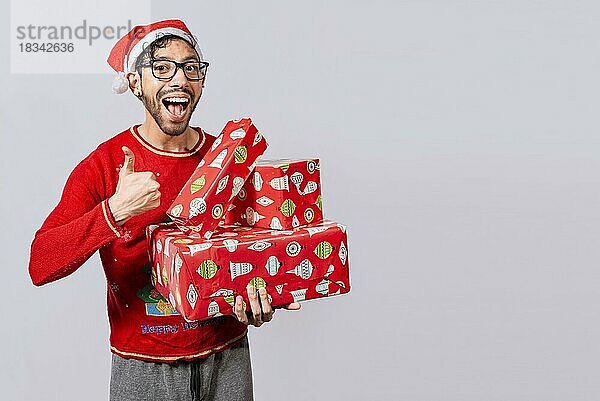 Lächelnder Mann in Weihnachtsmütze hält Weihnachtsgeschenkboxen und gibt Daumen hoch. Freundlicher Mann in Weihnachtskleidung hält Geschenkboxen  Weihnachtsmann hält Geschenkboxen und lächelt mit Daumen nach oben