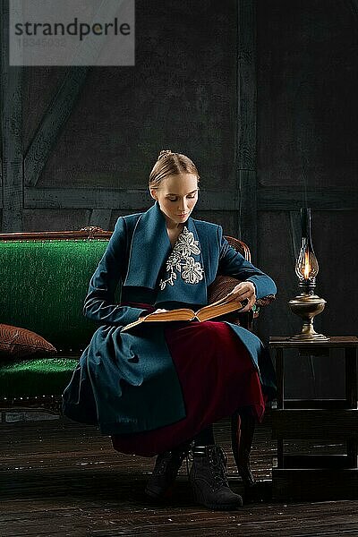 Attraktive Dame in altmodischer Kleidung liest altes Buch in der Nähe von Vintage Kerosinlampe