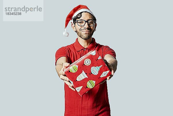 Lächelnder Mann mit Weihnachtsmütze bietet Ihnen eine Weihnachtsgeschenkbox an. Weihnachten Mann bietet ein Geschenk-Boxen isoliert. Handsome Mann in Weihnachten Kleidung zeigt ein Geschenk-Box