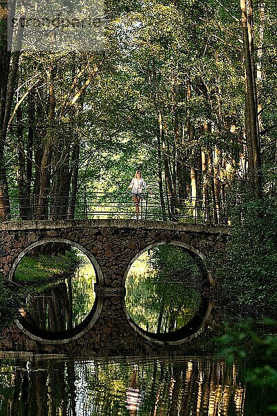 Vertikales Foto aus großer Entfernung von einem Mädchen auf einer Steinbrücke über einen kleinen Fluss im Park