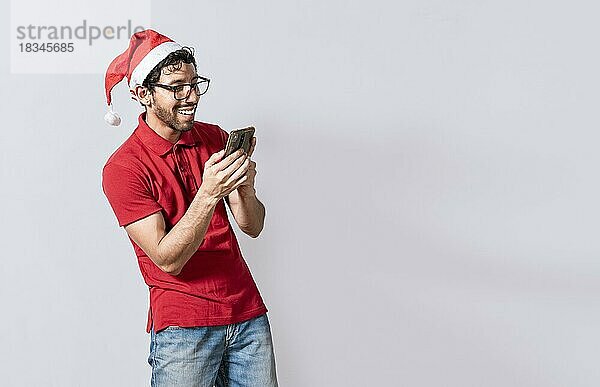 Lächelnder junger Mann in Weihnachtsmütze mit Handy isoliert. Glücklicher Mann in Weihnachtsmütze lächelnd am Handy isoliert. Menschen in weihnachtlichen Hut mit und lächelnd am Handy isoliert  Managua  Nicaragua  Mittelamerika
